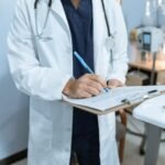 Médico de jaleco preenchendo atestado médico em prancheta, representando a emissão de um atestado médico rápido